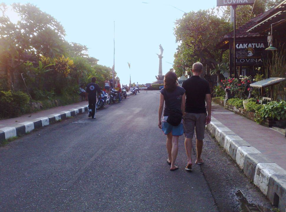 couple walking along the a quiet street in one of the towns of Lovina beach where in the distance you can see a dolphin statue