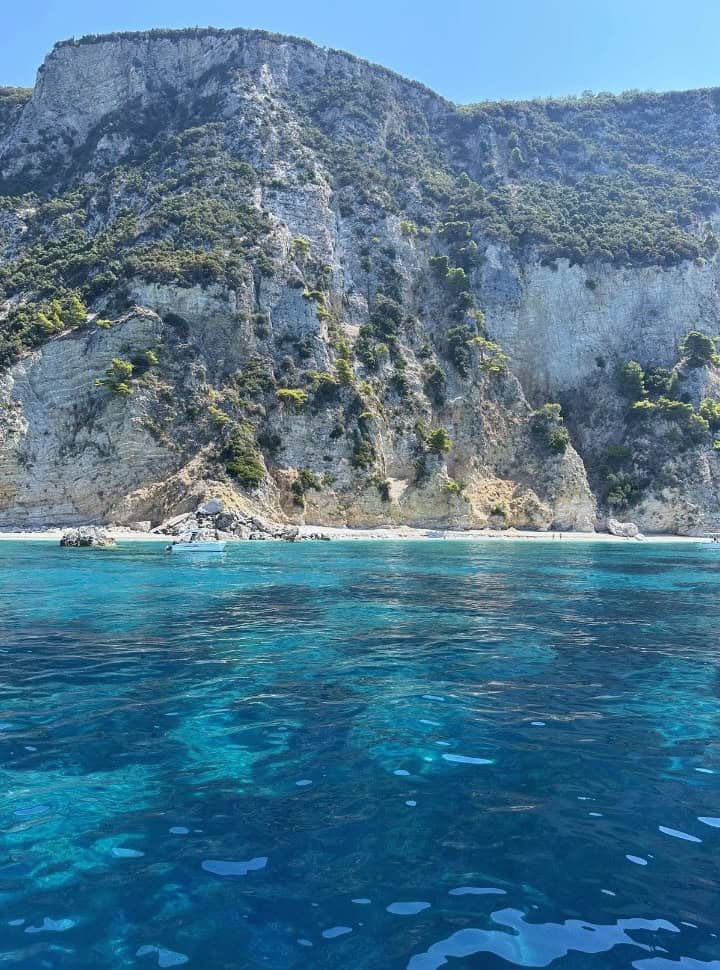 High white cliffs set at a small deserted beach with blue ocean, a small boat is seen along the Corfu coast