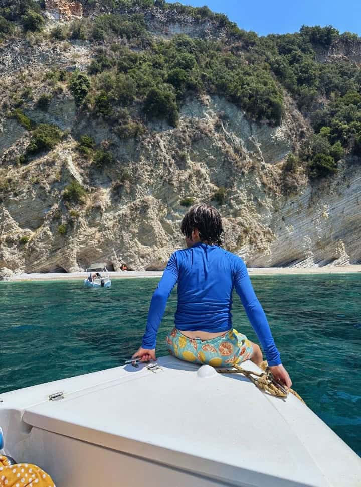 boy sitting on the tip of the boat looking at a white beach with a white cliff backdrop nearby Palaiokastritsa Corfu Greece