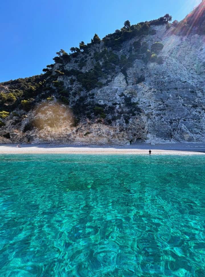 view from the water at Corfu Greece of a woman standing in a distance along the shore of a deserted white beach with turquoise water and white cliffs in the back