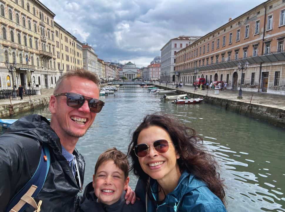 family of three posing for a selfie with in the background a canal with boats and on both sides old beautiful buildings in Trieste Slovenia
