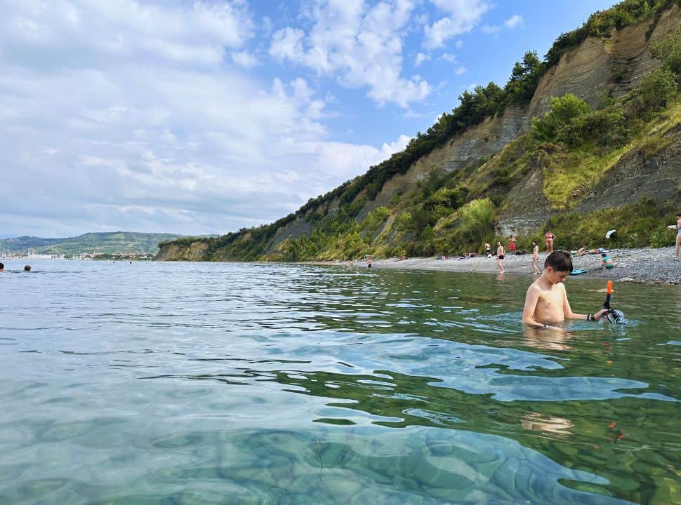 people swimming in the clear water at a pebbled beach set along the cliff along the coast of Slovenia