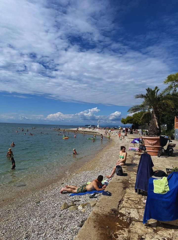 pebbled beach just along the promenade with calm clear water, lots of people enjoying the sunny weather in Izola Slovenia 