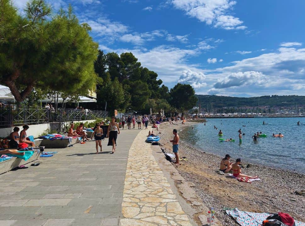 a small pebbled beach along a boulevard, where people are enjoying the sun while a lot of people are swimming in the water in the town Izola Slovenia