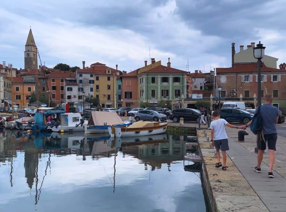 father and son walking towards Izola center from the harbor, a church tower stands between the colorful old buildings