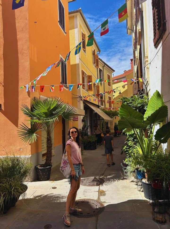 woman posing in a small street with orange and yellow buildings decorated with beautiful large plants and little flags in Izola Slovenia