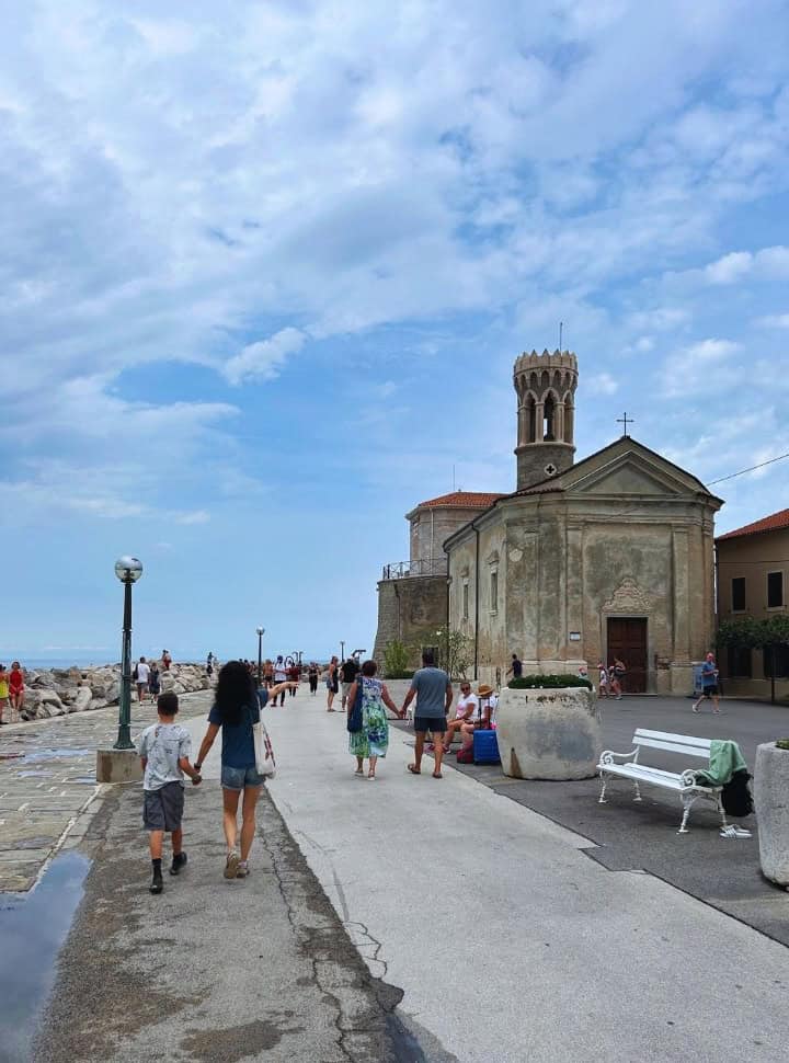 people walking along the promenade past a old church on one side and boulders and the ocean on the other in Piran Slovenia