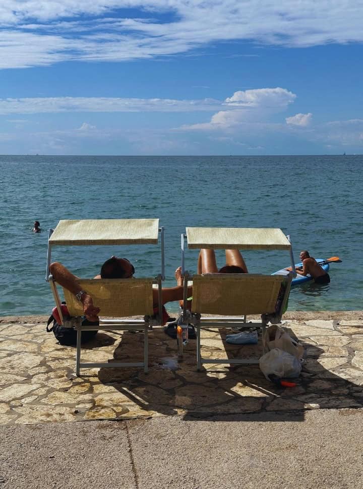 people relaxing on the sun loungers right on the waterfront, people swimming in the water in Izola, Sovenia